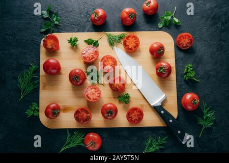 Top view of red sliced tomatoes on wooden chopping board. Sharp knife near. Green parsley and dill. Dark background. Preparing fresh vegetable salad Stock Photo