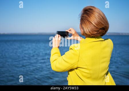 Stylish young woman in a bright yellow coat takes photos of the sea using a smartphone. Blank screen mobile phone. Close-up. Copy spase. Stock Photo