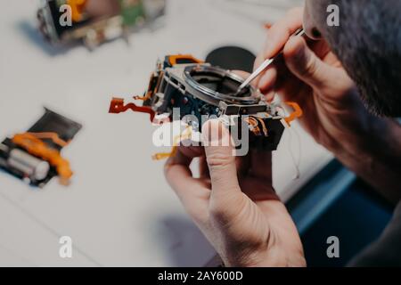 Repair process. Professional technician repairs broken digital camera, poses in service center or workshop, uses special tool. Maintenance and support Stock Photo