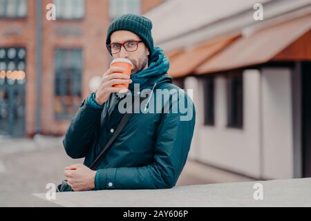 Outdoor shot of handsome hipster drinks takeawy coffee from disposable cup, dressed in headgear, jacket, wears spectacles, has srtoll outside, has str Stock Photo