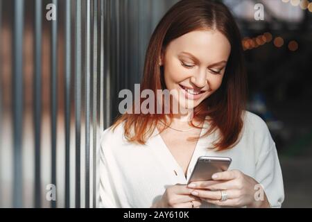 Pleased woman focused into screen of cell phone, checks email box, dressed in white clothes, sends feedback, connected to wireless internet, has brown Stock Photo