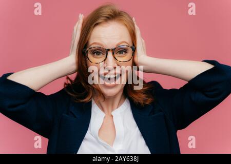 Photo of positive redhead female office worker keeps palms on ears, looks happily at camera, wears formal outfit, isolated over pink background. Peopl Stock Photo