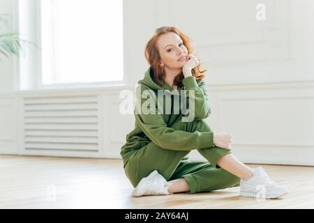 Staying in good shape. Pleasant looking red haired female gymnast with wavy hair, sits on floor in fitness hall, has flexible body, rests after doing Stock Photo