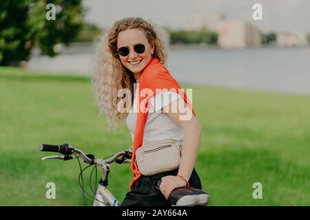 Happy curly haired blonde woman enjoys bicycle ride, has her own transport, leads healthy lifestyle, travels on bicycle around scenic nature views, dr Stock Photo