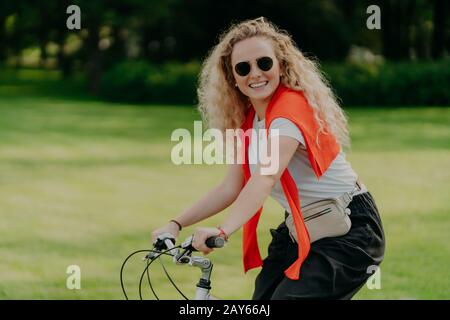 Photo of curly European woman keeps hands on handlebar of bicycle, travels outdoor in park, wears shades, casual t shirt and black trousers, smiles po Stock Photo