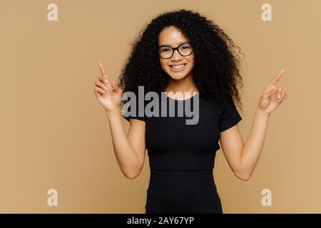 Positive joyful female model points above, shows something upwards, has friendly glad expression, slim body, wears casual black t shirt, isolated over Stock Photo