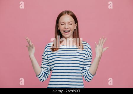 Waist up shot of overjoyed pretty woman gestures with hands, laughs positively, being in high spirit, dressed in sailor striped jumper, closes eyes sh Stock Photo