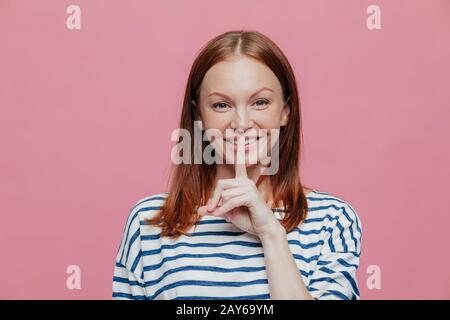 Photo of attractive woman with pleasant smile, holds fore finger over lips, being in good mood, tells nice secret, gossips or spreads rumors, wears st Stock Photo