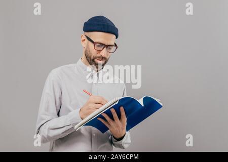 Serious male teacher writes down notes for next class, holds notepad, prepares interesting task for students, wears formal outfit, stands against grey Stock Photo