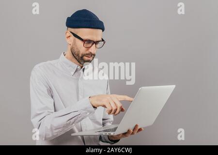 Professional male freelancer makes project work, points with index finger at screen of laptop computer, watches training webinar, holds modern gadget Stock Photo