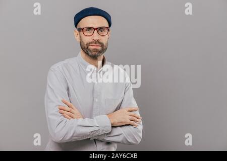 Photo of serious male keeps arms folded over chest, dressed in white formal shirt, fashionable hat, poses for making photography over grey wall with f Stock Photo
