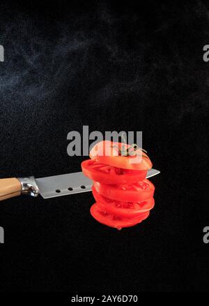 Flying red sliced tomato with a knife blade on a background of water dust in the backlight of a black background. Copy space, vertical image. Stock Photo