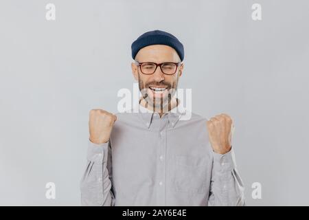 Gesture of success. Jubilant overjoyed unshaven man raises clenched fists, wears spectacles and formal shirt, celebrates his triumph, closes eyes from Stock Photo