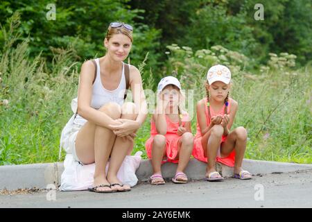 Mother and two children sitting on the curb of the road waiting for the bus Stock Photo