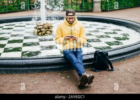 Fashionable traveler in black cap, glasses, jeans and yellow jacket sitting near fountain in park playing games on his smartphone having rest after lo Stock Photo