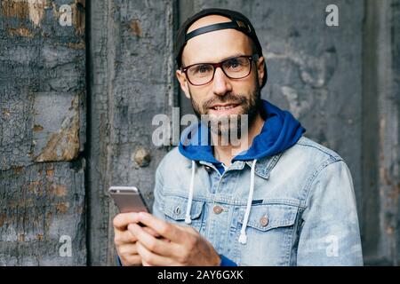 Close-up portrait of bearded blue-eyed man in stylish cap and denim anorak holding mobile phone surfing the Internet cheking his emails and communicat Stock Photo