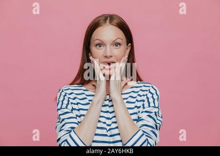 Beautiful dark haired amazed woman touches cheeks with both hands, dressed in striped clothes, looks surprisingly at camera, poses against pink backgr Stock Photo