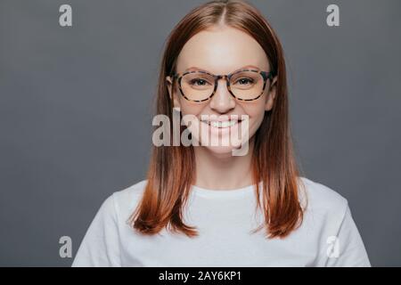 Headshot of pleasant looking dark haired young woman wears transparent glasses, dressed in casual clothes, has healthy skin, positive look, shows whit Stock Photo