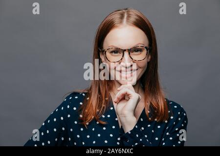 Happy delighted young woman keeps one hand under chin, smiles happily, wears transparent glasses, elegant clohtes, has eyes full of happiness, isolate Stock Photo