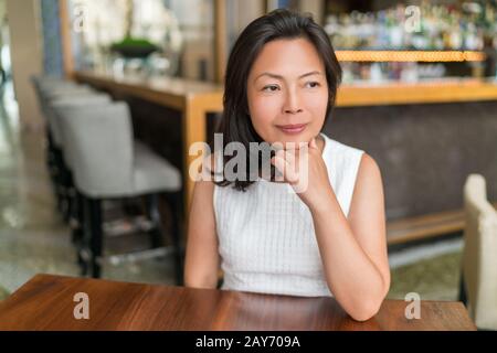 Elegant Asian middle-aged businesswoman pensive portrait. Beautiful mature Chinese business woman relaxing in restaurant bar urban stylish living in Shanghai, China. Mindfulness concept. Stock Photo