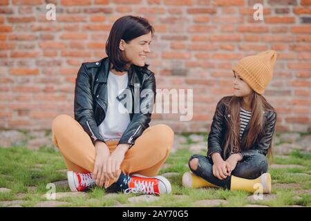 Shot of pleased realxed two sisters pose crossed legs, sit on lawn, pose against brick wall background, have happy facial expressions. Little kid in l Stock Photo