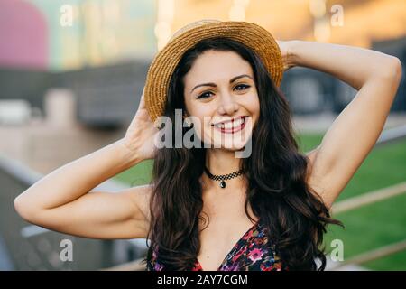 Happy brunette female with pleasant smile wearing hat keeping hand behind head posing outdoors. Pretty woman with happy expression relaxing at big cit Stock Photo