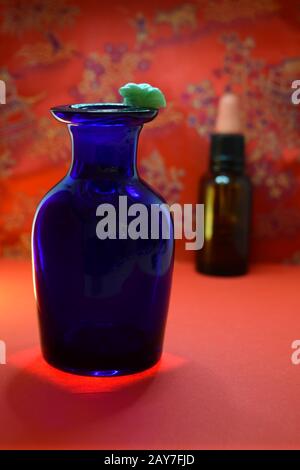 Blue glass bottle lit from below  with small jade frog on rim, brown glass bottle with rubber stopper in background against a red Chinese pattern. Stock Photo