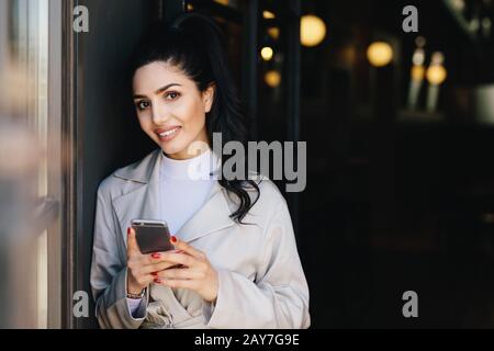 Smiling pretty brunette woman with pony tail having red manicure dressed formally holding smartphone doing online shopping while waiting for train. Bu Stock Photo