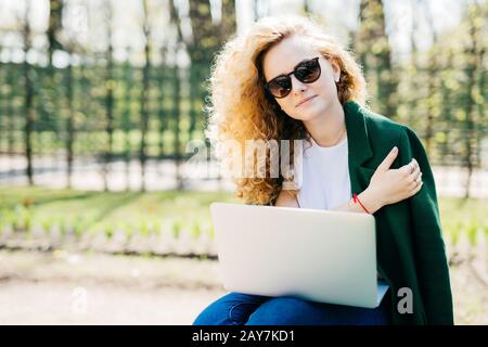 Portrait of cute young woman with curly blonde hair wearing sunglasses, white T-shirt and green jacket holding laptop on knees working at her future p