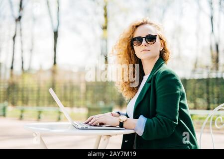 Sideways portrait of cute blonde female in elegant clothes typing something on her laptop looking aside with pensive expression dreaming about her fut Stock Photo