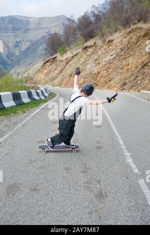 Longboarder on longboard in overalls helmet and gloves performs a stand-up slide at speed while on a mountain road serpentine in Stock Photo