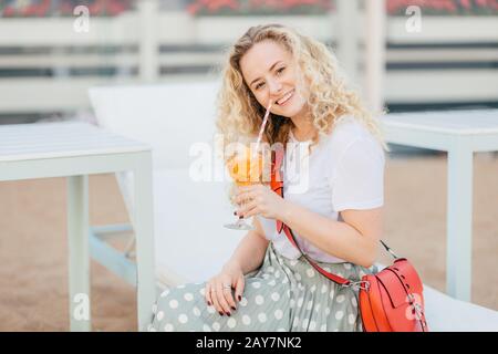 Joyful female model with friendly smile, healthy pure skin, holds glass of cocktail, wears stylish clothes, recreats on beach during summer vacation, Stock Photo