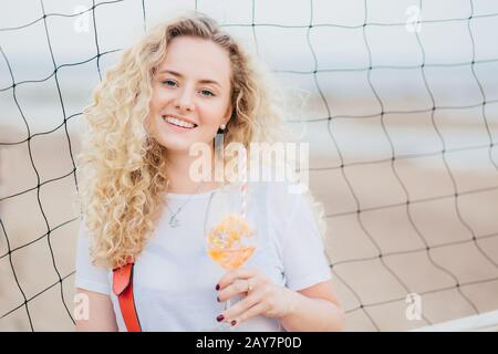 Optimistic young female model with curly light hair, dressed in casual white t shirt, drinks fresh cocktail, enjoys good rest at beach, stands near te Stock Photo