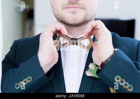 A sturdy newlywed with a beard in a suit adjusts the wooden bow tie on the collar with his white shirt. Stock Photo