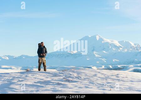A traveler with a large backpack on his shoulders Stand on a snow-capped hill against the blue sky and the sleeping Elbrus volca Stock Photo