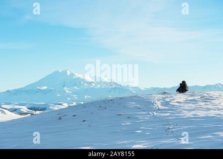 A traveler with a large backpack on his shoulders sits on a snow-capped hill against the blue sky and the sleeping Elbrus volcan Stock Photo
