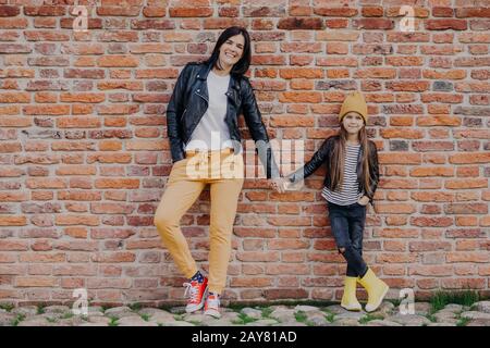 Motherhood, relationship and style concept. Fashionable young mother in leather jacket and sport shoes holds hand of her little daughter dressed in st Stock Photo