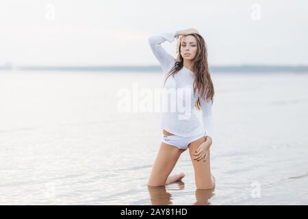 Outdooor view of good looking brunette female with make up, dressed in white bikini, poses at camera near calm sea background, has fit body shape, fee Stock Photo