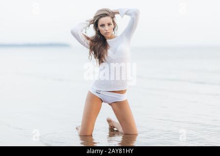 Portrait of attractive female with make up, dressed in white clothes, stands on knees at sandy beach near ocean view, has confident expression, feeles Stock Photo