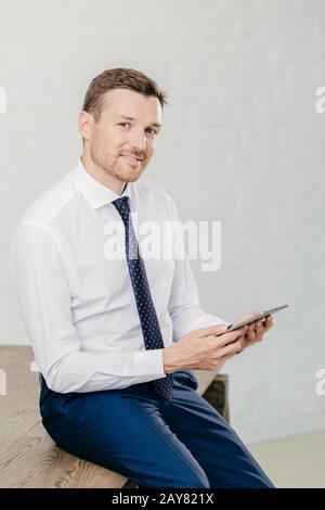 Confident pleased young male entrepreneur in luxury formal clothes, reads recieved text message from colleague on modern tablet computer, looks joyful Stock Photo