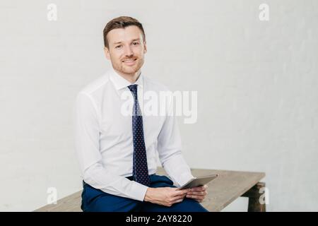Indoor shot of male financier with positive expression wears formal white shirt with tie, holds modern tablet computer in hands, isolated over white b Stock Photo
