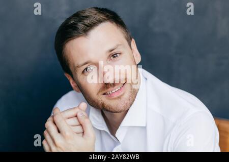 Attractive unshaven male office worker has break after work, looks joyfully at camera, going to have good rest, wears elegant white shirt, poses indoo Stock Photo