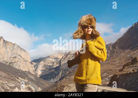 A girl photographer in sunglasses and a big fur hat and a yellow knitted sweater stands against the background of high rocks in Stock Photo