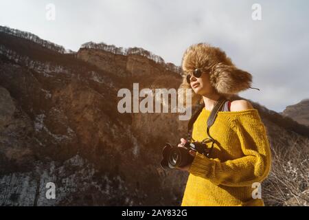 A girl photographer in sunglasses and a big fur hat and a yellow knitted sweater stands against the background of high rocks in Stock Photo