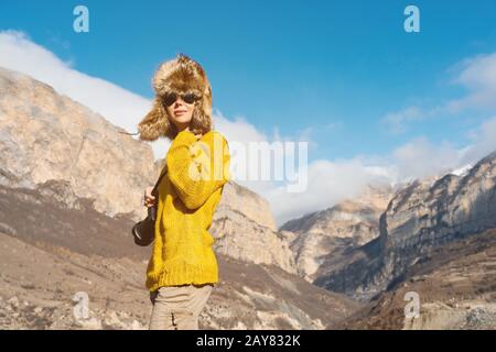 A girl photographer in sunglasses and a big fur hat and a yellow knitted sweater stands against the background of high rocks in Stock Photo