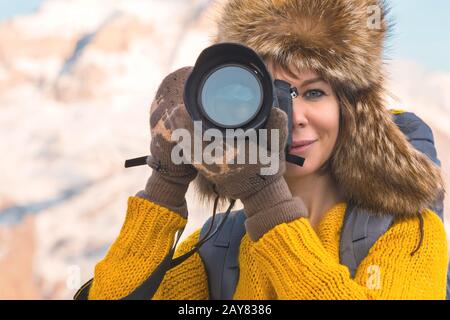 Portrait of a sweet tourist girl in a big fur hat takes pictures on her digital camera in the mountains. Stock Photo