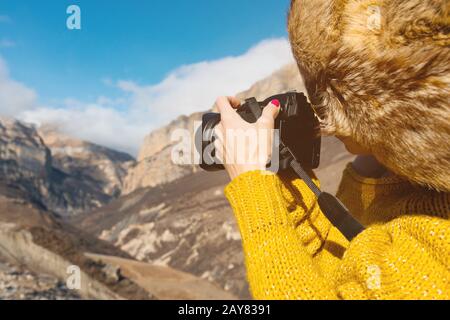 Close up A girl photographer in a fur hat and a yellow sweater in the mountains takes pictures on her digital camera. Stock Photo