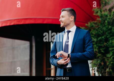 Horizontal shot of satisfied businessman in formal clothing, looks at watch, waits for partner near cafe, going to discuss details of collaboration, h Stock Photo