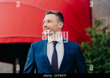 Cheerful manager dressed in formal outfit, has pleasant smile on face, being in good mood after successfully signed contract, poses outdoor against re Stock Photo