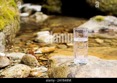 A transparent glass glass with mineral mountain river water stands on a stone beside the mountain river creek Stock Photo